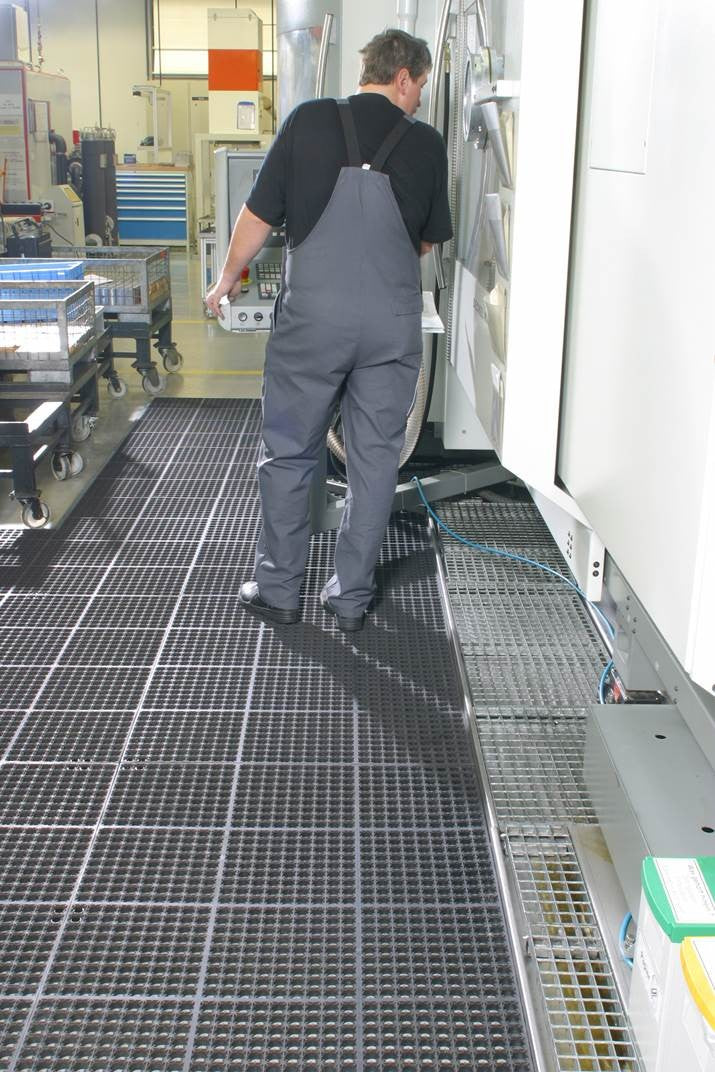Dark anti-fatigue floor mat with square grid tread, used in a production aisle